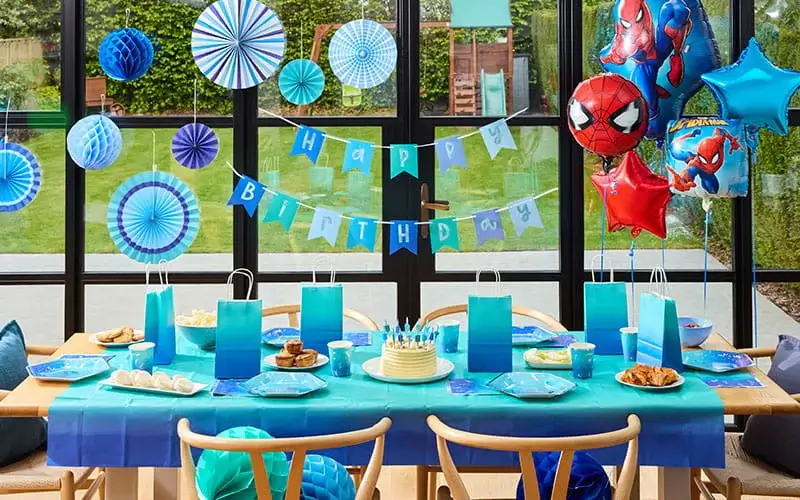 Blue birthday party table with a birthday cake, gift bags, themed decorations, hanging paper fans, and superhero balloons.